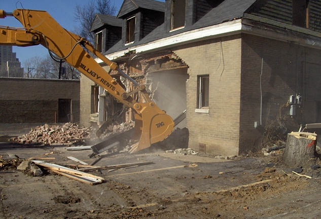 A yellow steam shovel begins demolition on a building.