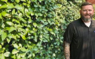 Pennington in black polo standing in front foliage smiling at camera
