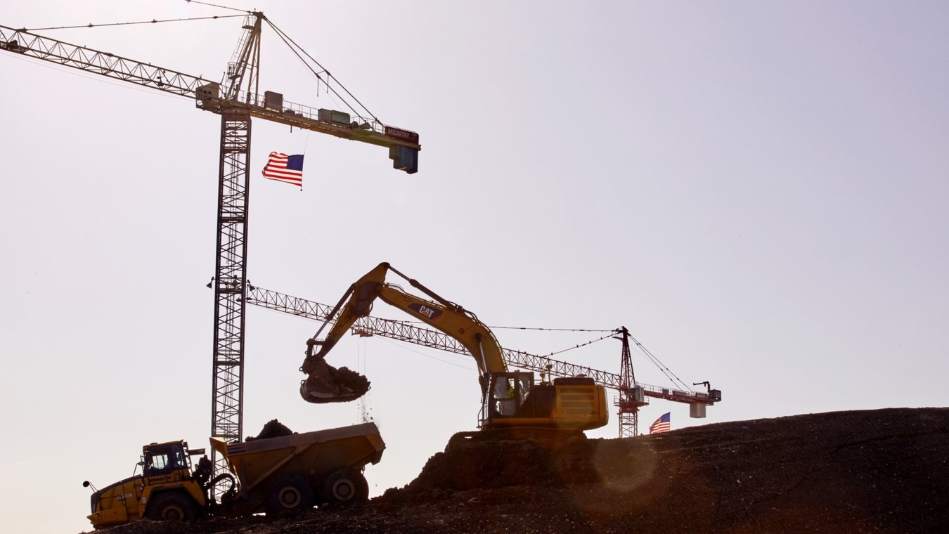 Foreground CAT excavator placing dirt in dump truck two and two cranes standing in background