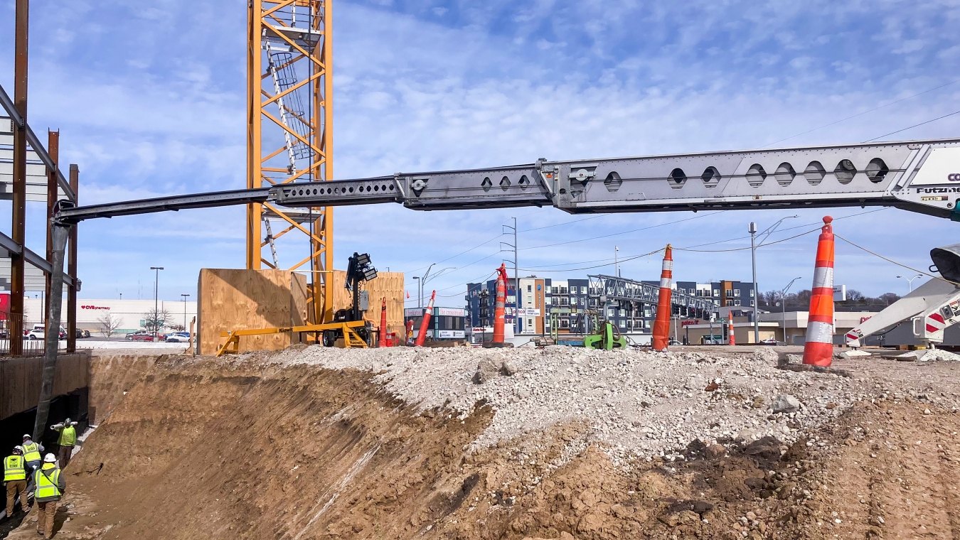 Workers in pit pouring concrete next to building foundation with crane tower in background