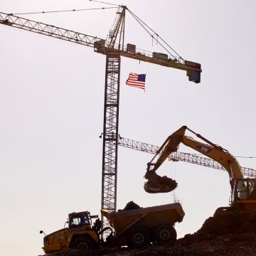 Foreground CAT excavator placing dirt in dump truck two and two cranes standing in background