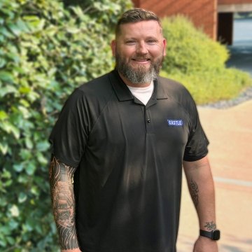 Pennington in black polo standing in front foliage smiling at camera